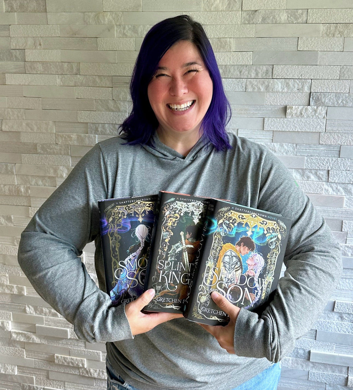 Author Gretchen Powell Fox smiling and holding the three Shattered Crown hardcovers fanned out